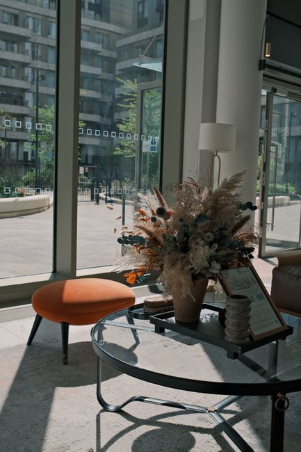 A large floral arrangement featuring a variety of dried and preserved flowers in neutral tones including beige, white, and light brown, with hints of muted orange and pink. The bouquet is placed in a 