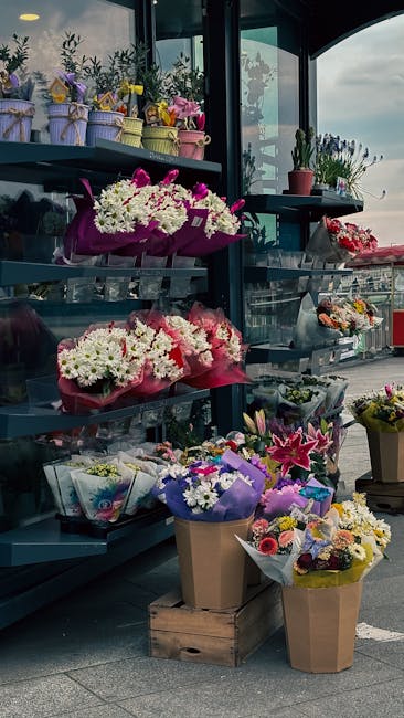 An outdoor display of floral arrangements on metal shelves, featuring a variety of fresh flowers including white daisies, pink lilies, and purple statice, wrapped in colorful paper and cellophane. The