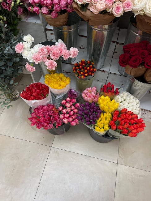 A display of various fresh flowers arranged in small bouquets and placed on the floor in front of larger flower arrangements in metal buckets at a florist shop. The smaller bouquets include vibrant re
