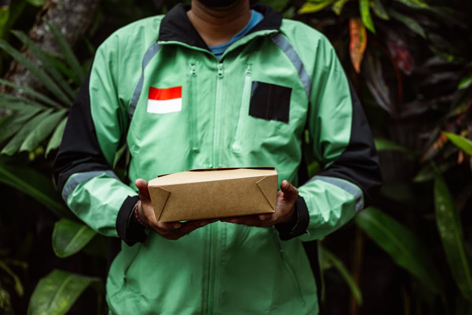 A delivery person wearing a bright green jacket with black accents, featuring a small Indonesian flag patch on the chest, is holding a rectangular brown cardboard gift box with folded corners, positio