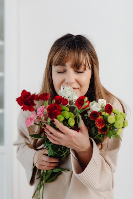 A woman with light brown hair and bangs, dressed in a beige sweater, holds a vibrant, fresh floral bouquet close to her face with her eyes closed, smiling gently. The bouquet features a mix of red car