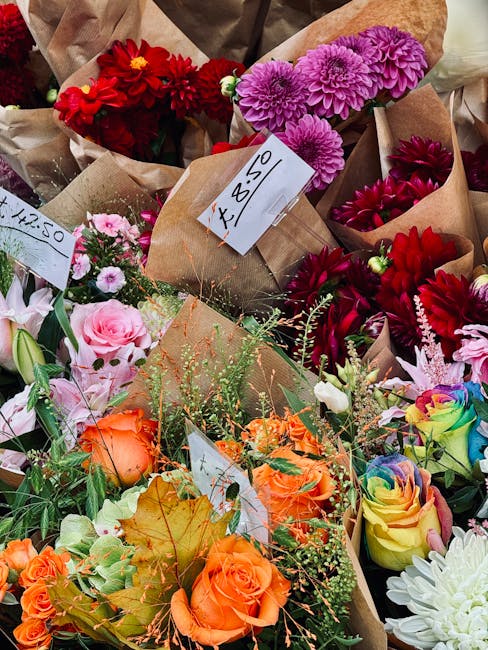 A vibrant assortment of fresh flowers arranged in multiple small brown paper bouquets, featuring a variety of species and colors including bright red, deep pink, lavender, orange, yellow, and white. T